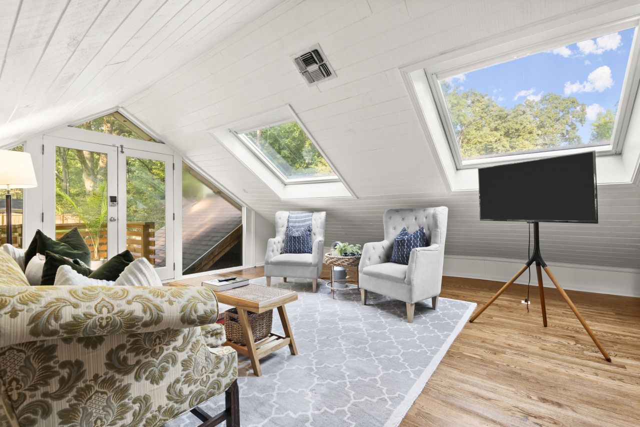 A room with a sloped ceiling, skylights, a sofa, armchairs, a TV on a tripod stand, and a small table with a lamp.