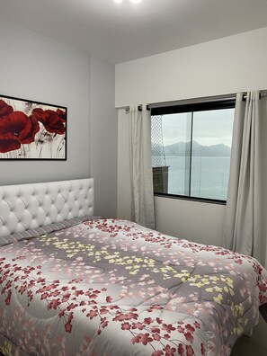 A bedroom with a tufted headboard, a floral bedspread, a large window with curtains, and a painting of red flowers on the wall. - Apartment with spectacular views, Porto Real Resort (Mangaratiba)