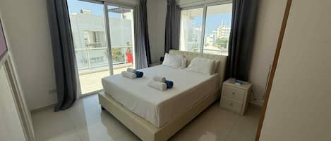 A modern hotel room with a large bed, a bedside table, and a view of the city through large windows.