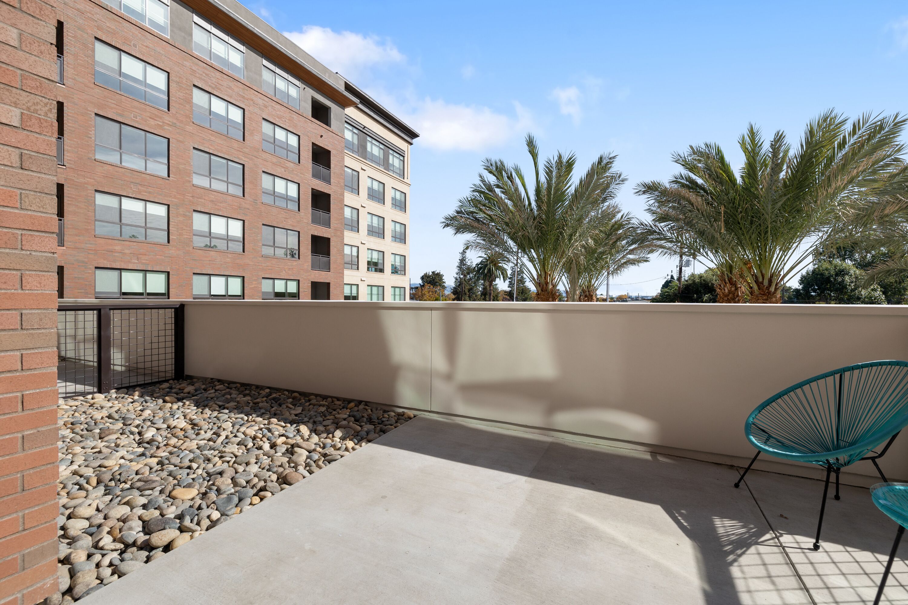 A rooftop patio with a concrete floor, a brick wall, and a metal railing. There are two turquoise chairs and a palm tree in the background.