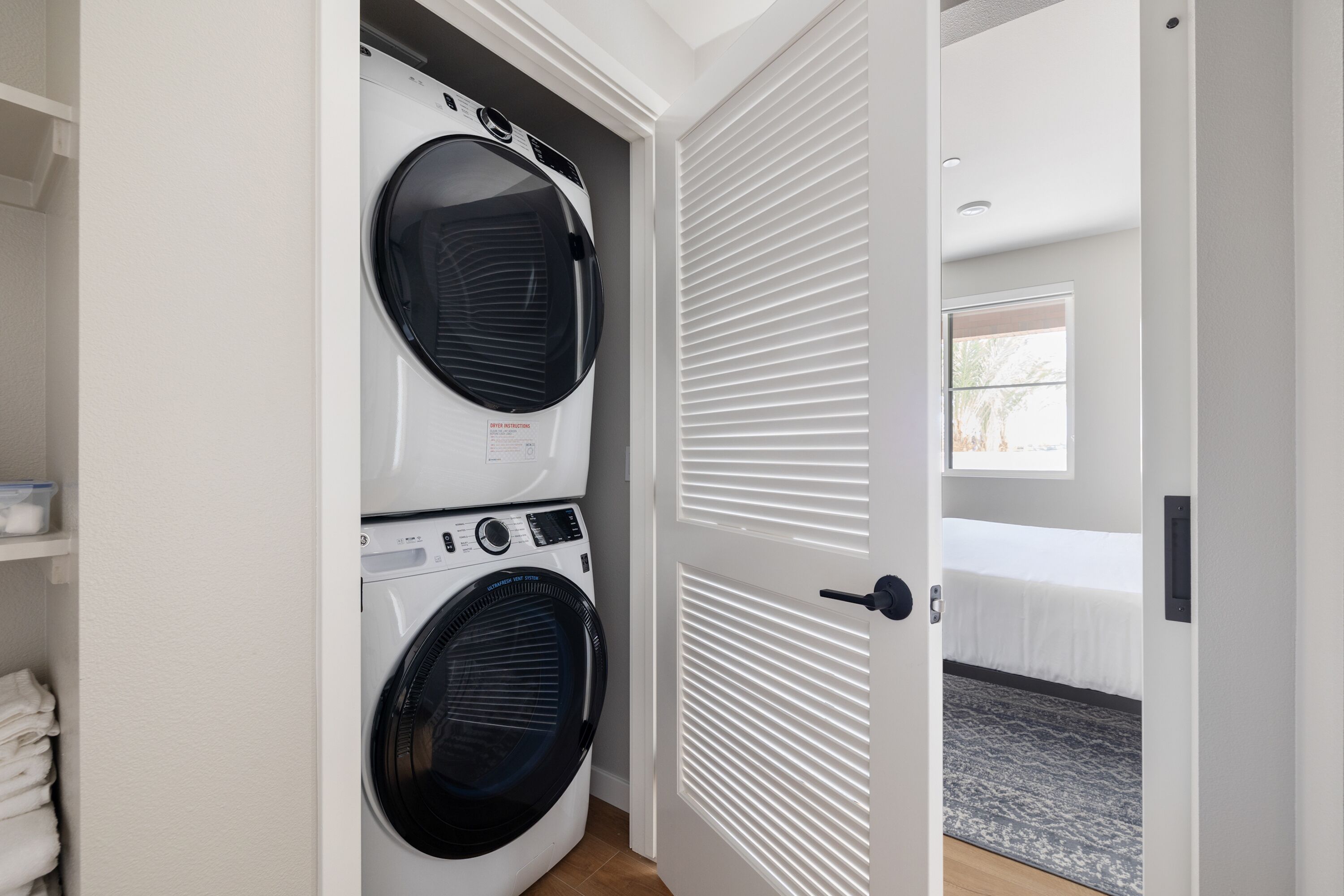 Ein Waschraum mit zwei übereinander gestapelten Waschmaschinen mit Frontlader, einer weißen Tür zu einem Schlafzimmer mit Bett und Fenster.
