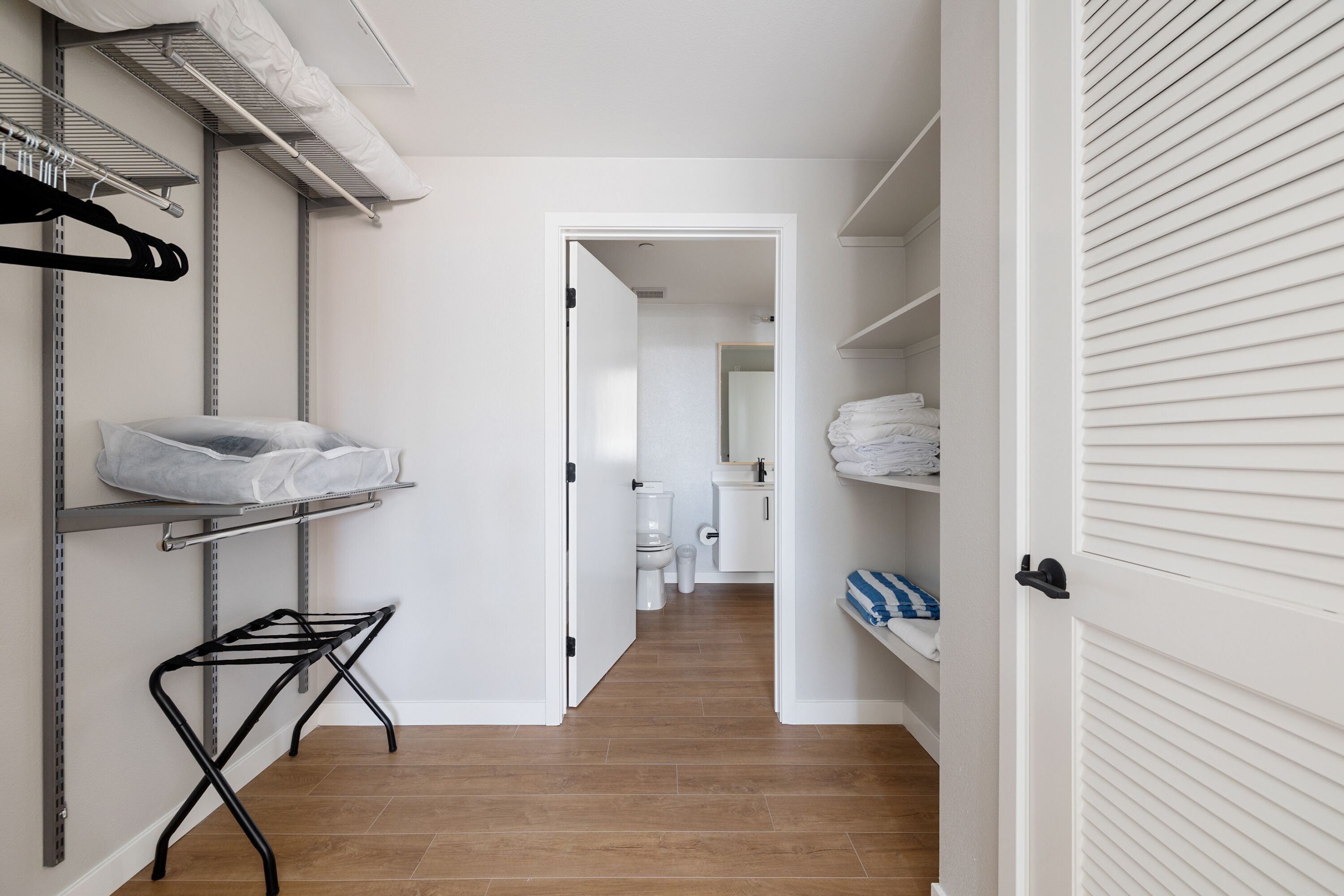 A narrow hallway with a metal rack holding folded clothes, a drying rack, and a white door leading to a bathroom with a toilet and sink.