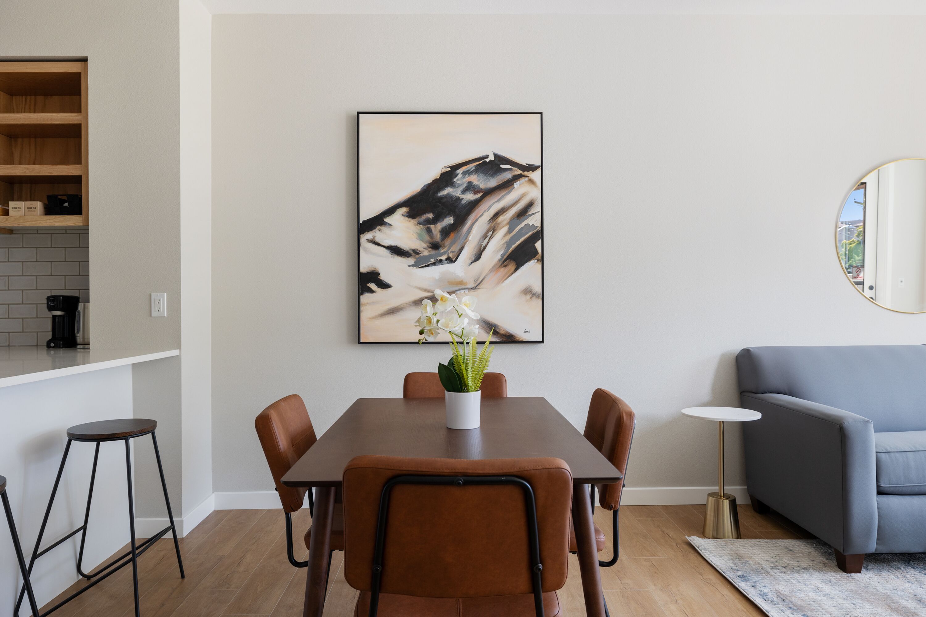 Un área de comedor con una mesa rectangular, seis sillas, un jarrón con flores, un cuadro en la pared, un espejo redondo, un sofá y una pequeña mesa redonda.