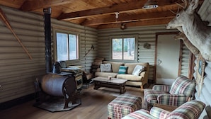 Interior de cabana de tronco com teto de madeira, lareira, mobiliário com estampa xadrez e cabeça de veado montada na parede.