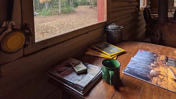 Interior de uma cabana com uma janela grande, uma mesa com livros e uma caneca verde, e uma parede de madeira.