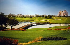 A golf course with a bridge over a pond, surrounded by greenery and buildings in the background.