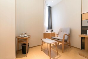 A small, modern room with a wooden floor, a grey upholstered chair, a small table with a white cushion, and a window with curtains. - Ji Hotel (Shanghai)