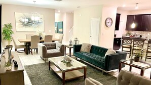 A spacious living room with a green sofa, beige armchairs, and a glass coffee table. There is a dining area with a table and chairs, and a kitchen area with dark cabinets and a microwave. - Modern 5BR/5BA Vacation Home with Private Pool CG1593 (Davenport)