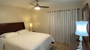 Une chambre d’hôtel avec un lit, une table de chevet, une lampe, un ventilateur de plafond et une fenêtre avec des rideaux.