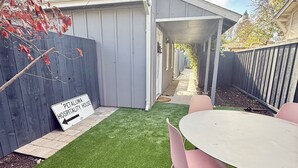 A backyard with artificial grass, a patio table with chairs, a wooden fence, and a sign indicating a hospitality house.