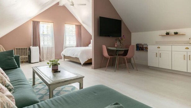 A cozy, well-lit room with a bed, a dining table with chairs, a sofa, a TV mounted on the wall, and a kitchenette area.