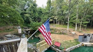 En träveranda med en liten damm, en amerikansk flagga och en träkonstruktion i ett skogsområde.