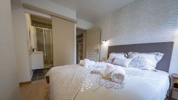Una cama bien hecha con una colcha blanca y cojines decorativos, una mesita de noche de madera, y un baño con ducha e inodoro a la vista.