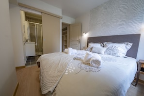 Un lit bien fait, avec une couette blanche et des coussins décoratifs, une table de chevet en bois et une salle de bain où l’on peut voir une douche et des toilettes.