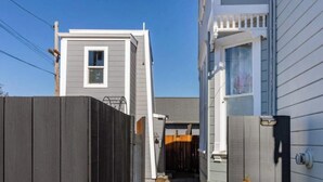 Ein Wohnhaus mit einem schmalen Weg, der zu einer Garage führt. Das Haus hat eine Fassade in Weiß und Grau, ein kleines Fenster und eine Tür mit Glasfenster.