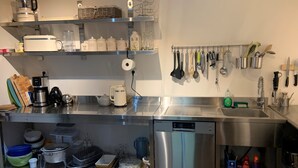 Una cocina moderna con electrodomésticos de acero inoxidable, incluyendo una cafetera, un hervidor y un lavavajillas. Hay estantes con recipientes de almacenamiento y un escurreplatos con utensilios de cocina.