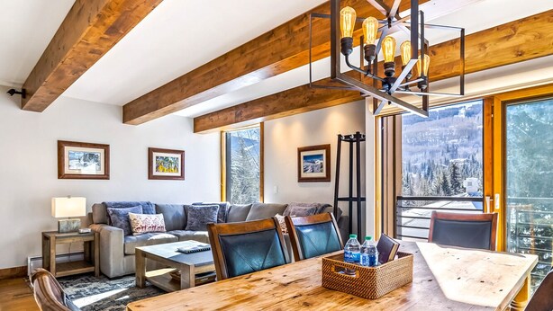 Un salon chaleureux avec un plafond en bois, un ensemble de canapés, une table à manger et une grande fenêtre donnant sur les montagnes.