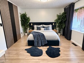 A modern bedroom with a bed, two nightstands, a headboard, a window with curtains, and two potted plants.