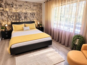 A modern bedroom with a large bed, a nightstand with a lamp, a potted plant, and a view of a forest through sheer curtains.