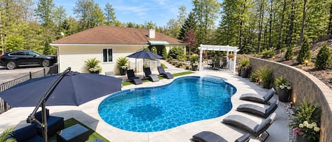 Una piscina con sillones de descanso y una pérgola, rodeada de un jardín ajardinado y una casa.