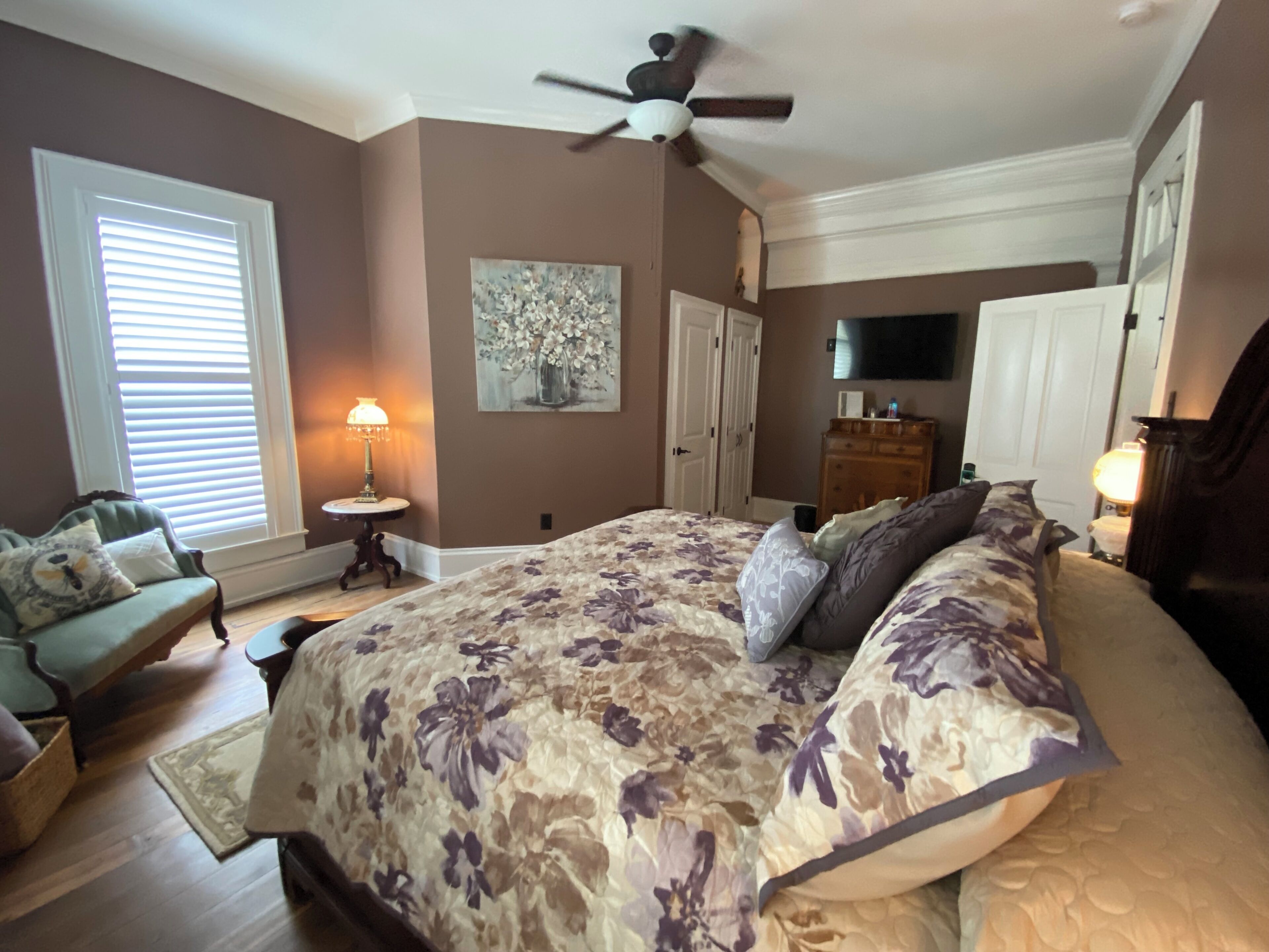A bedroom with a large bed, a ceiling fan, a TV, and a painting on the wall.