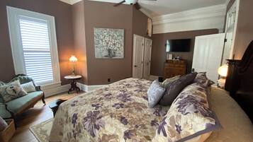 A bedroom with a large bed, a ceiling fan, a TV, and a painting on the wall.