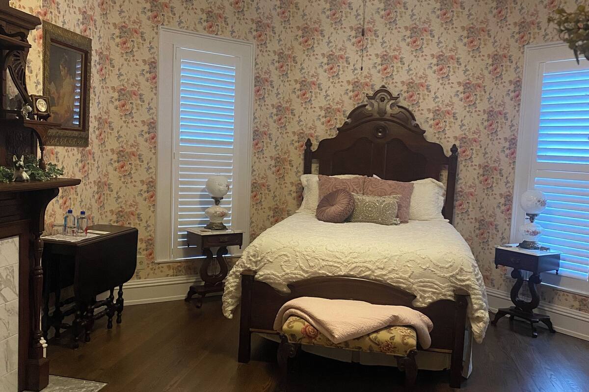 Een slaapkamer met een houten bed, bloemenbehang, een plafondventilator en een groot vloerkleed.