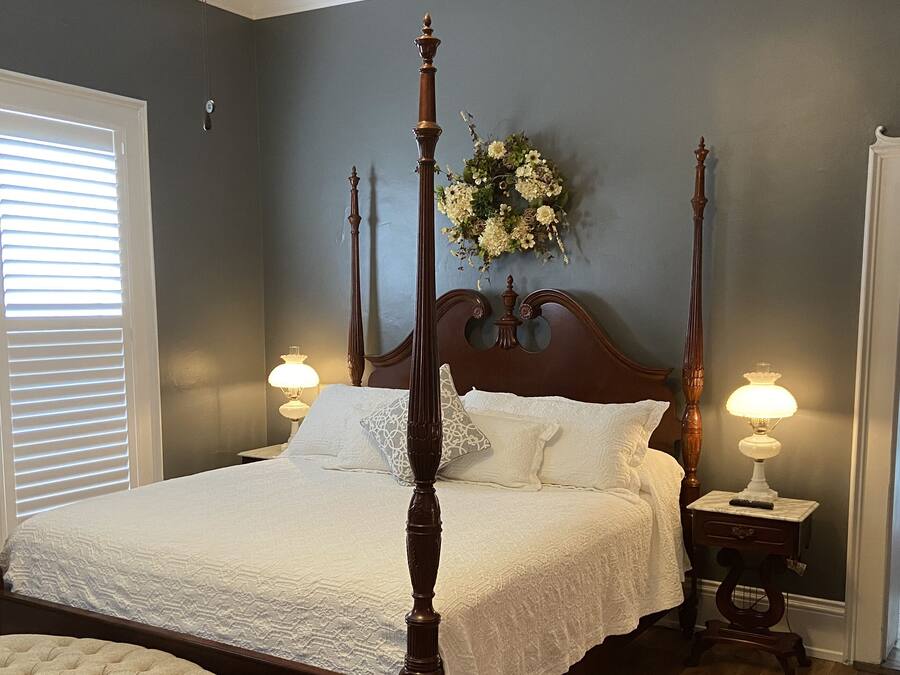 A four-poster bed with white linens, two bedside tables with lamps, and a floral arrangement on the wall.