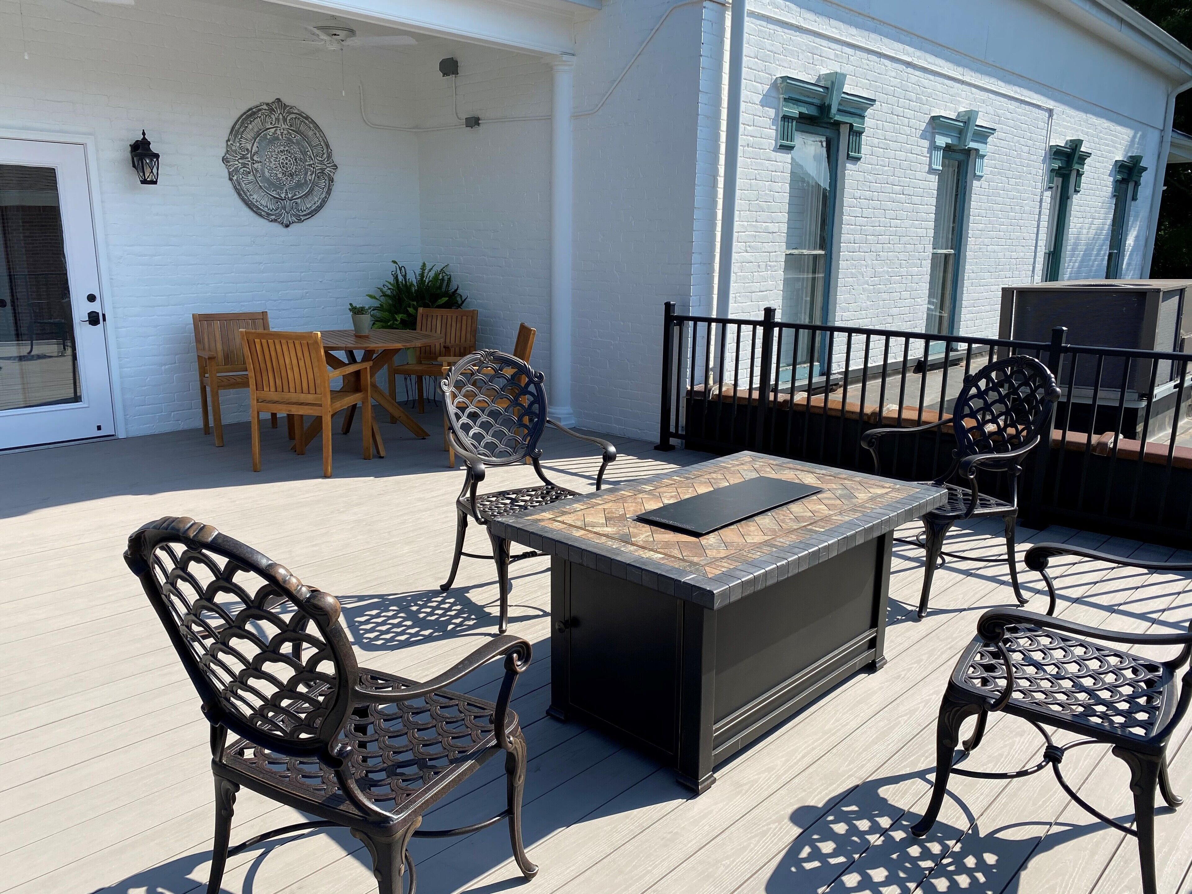 Een overdekt terras met een houten tafel en stoelen, een decoratieve muur en een wit gebouw met ramen.