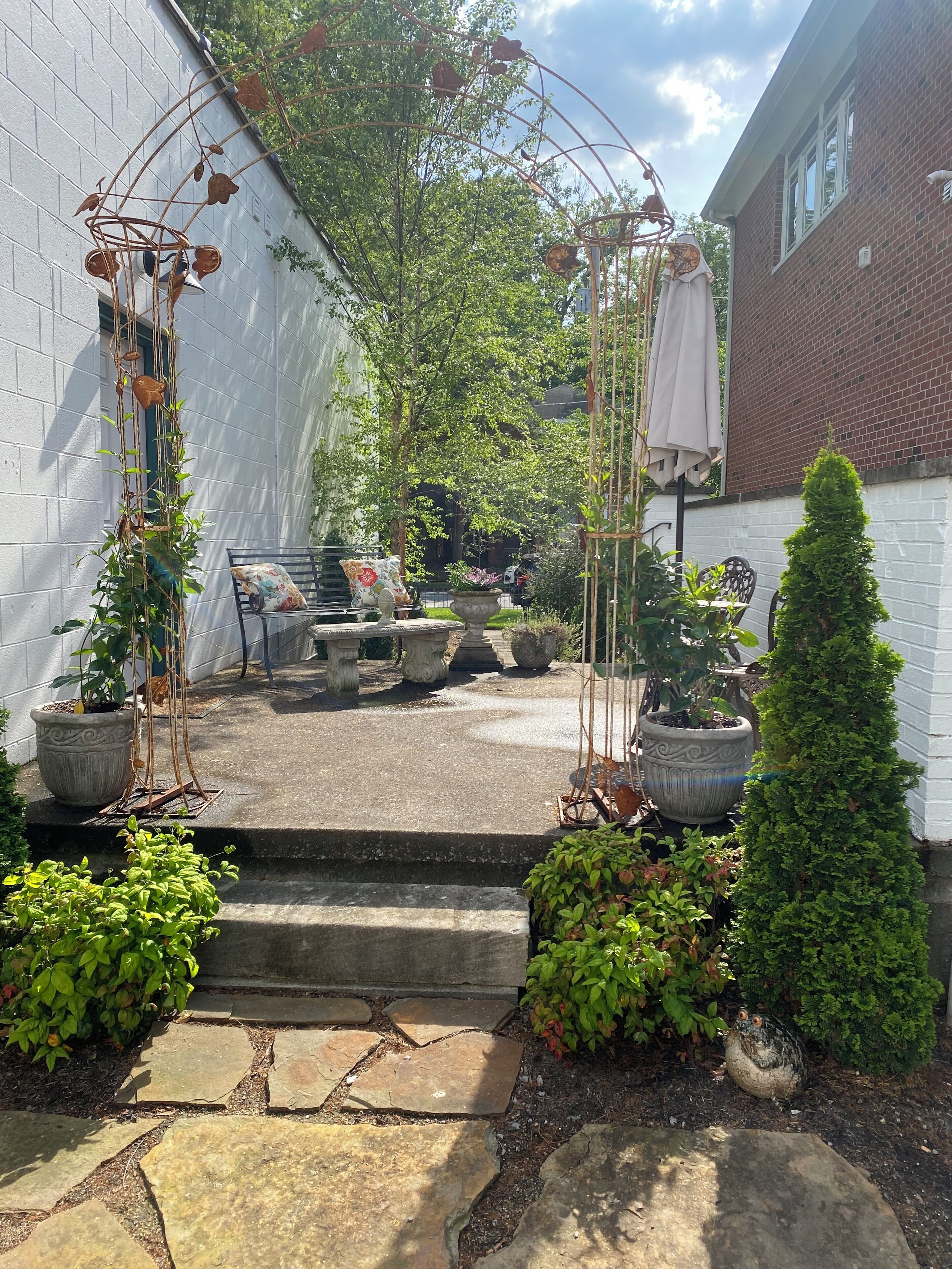 Un patio con un sendero de piedra que lleva a una zona de estar, rodeado de plantas en macetas y un arco de metal decorativo.