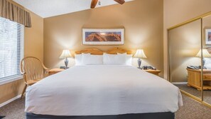 Quarto de hotel com uma cama grande, duas luminárias laterais, uma cadeira de madeira, um quadro na parede e um espelho no lado direito.