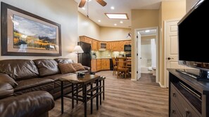 A living room with a leather couch, a coffee table, and a TV. There is a kitchen in the background with wooden cabinets and a dining area.