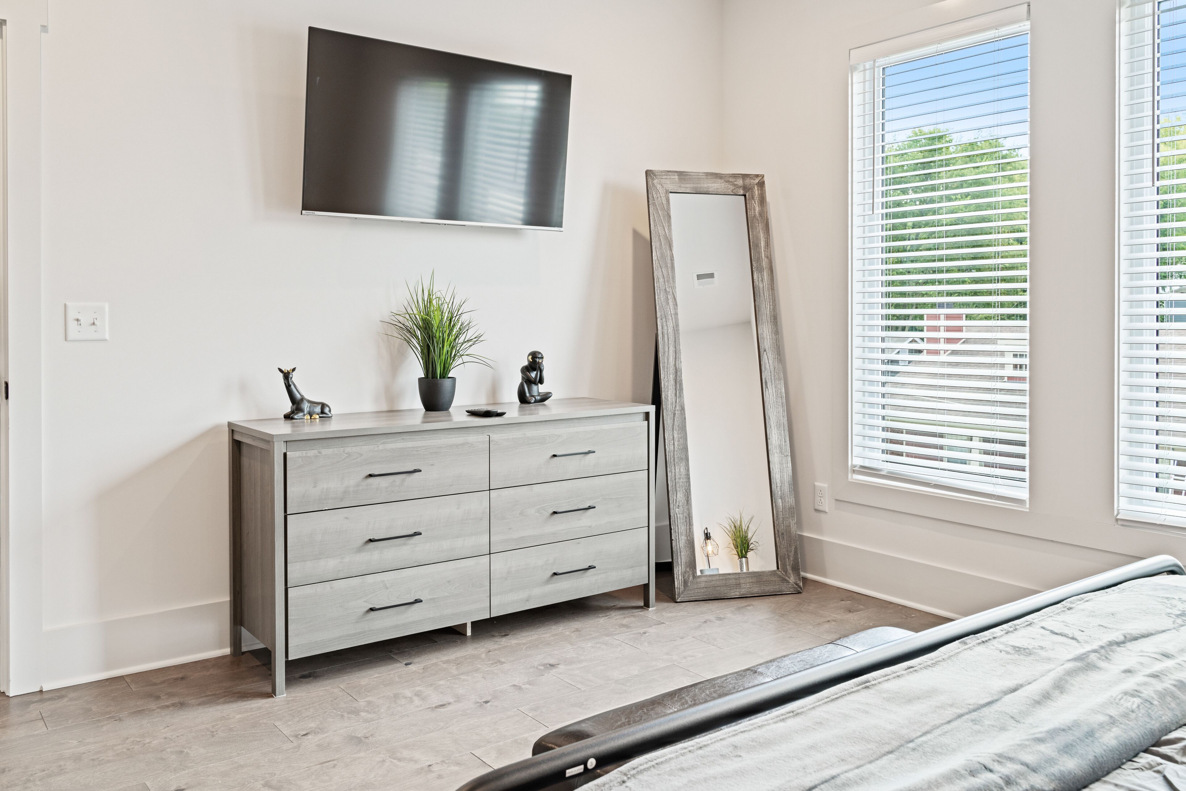 Une chambre à coucher comprenant un lit, une commode, un téléviseur, un grand miroir et une fenêtre avec des stores.