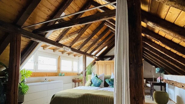 An attic bedroom with a sloped ceiling, wooden beams, a bed with green bedding, a window, and a plant. - Historic vacation home on the Main, 110 sqm, large terrace, modern and renovated (Mainz-Kostheim)