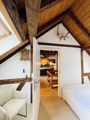 Ein gemütliches Schlafzimmer im Dachgeschoss mit schrägem Dach, einem Holzbalken, einem Bett, einem Sessel und einem kleinen Tisch.