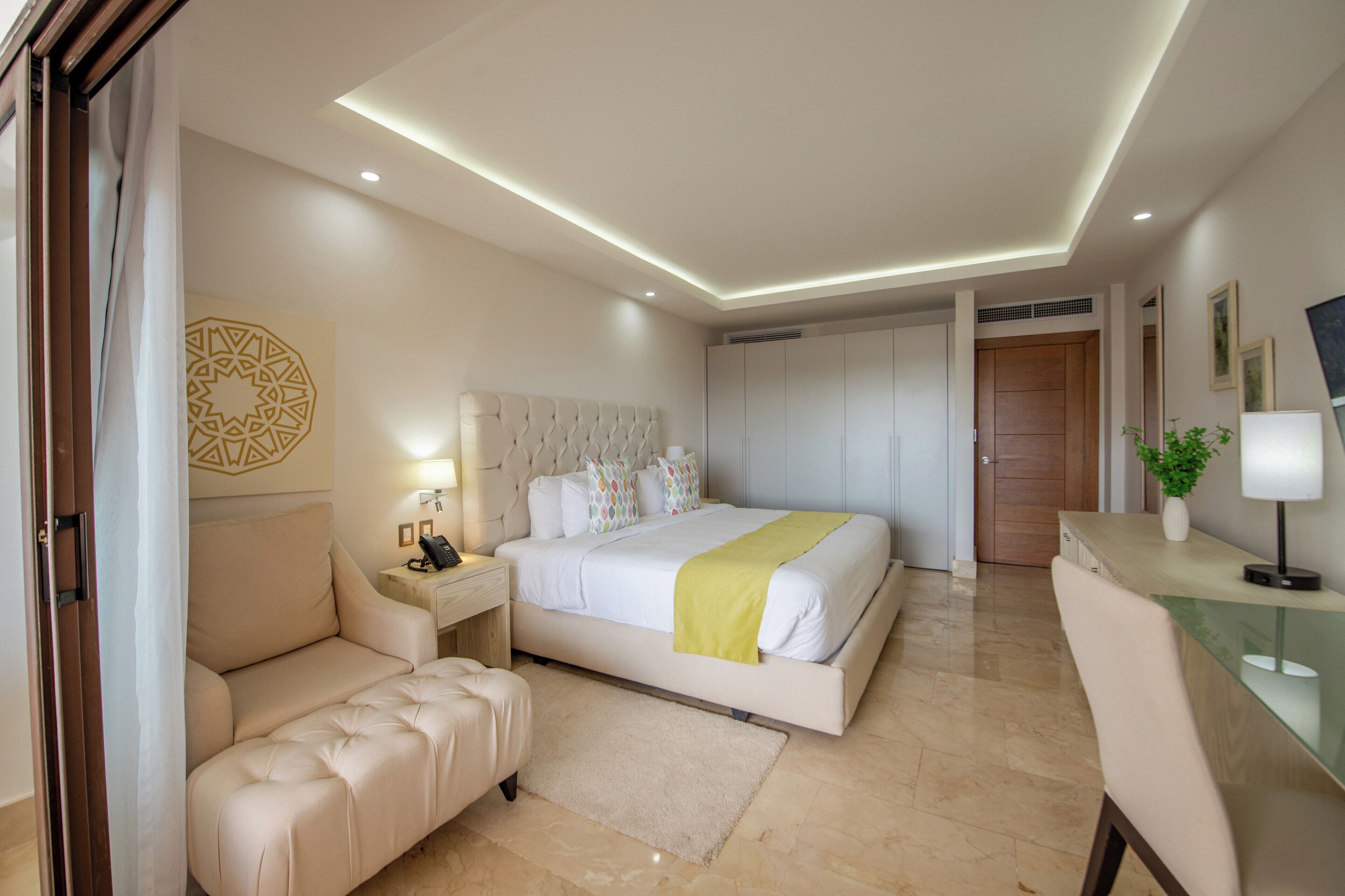 Une chambre d’hôtel moderne dotée d’un grand lit, d’un coin salon avec un canapé et un pouf, d’un bureau avec une lampe et d’un grand miroir.
