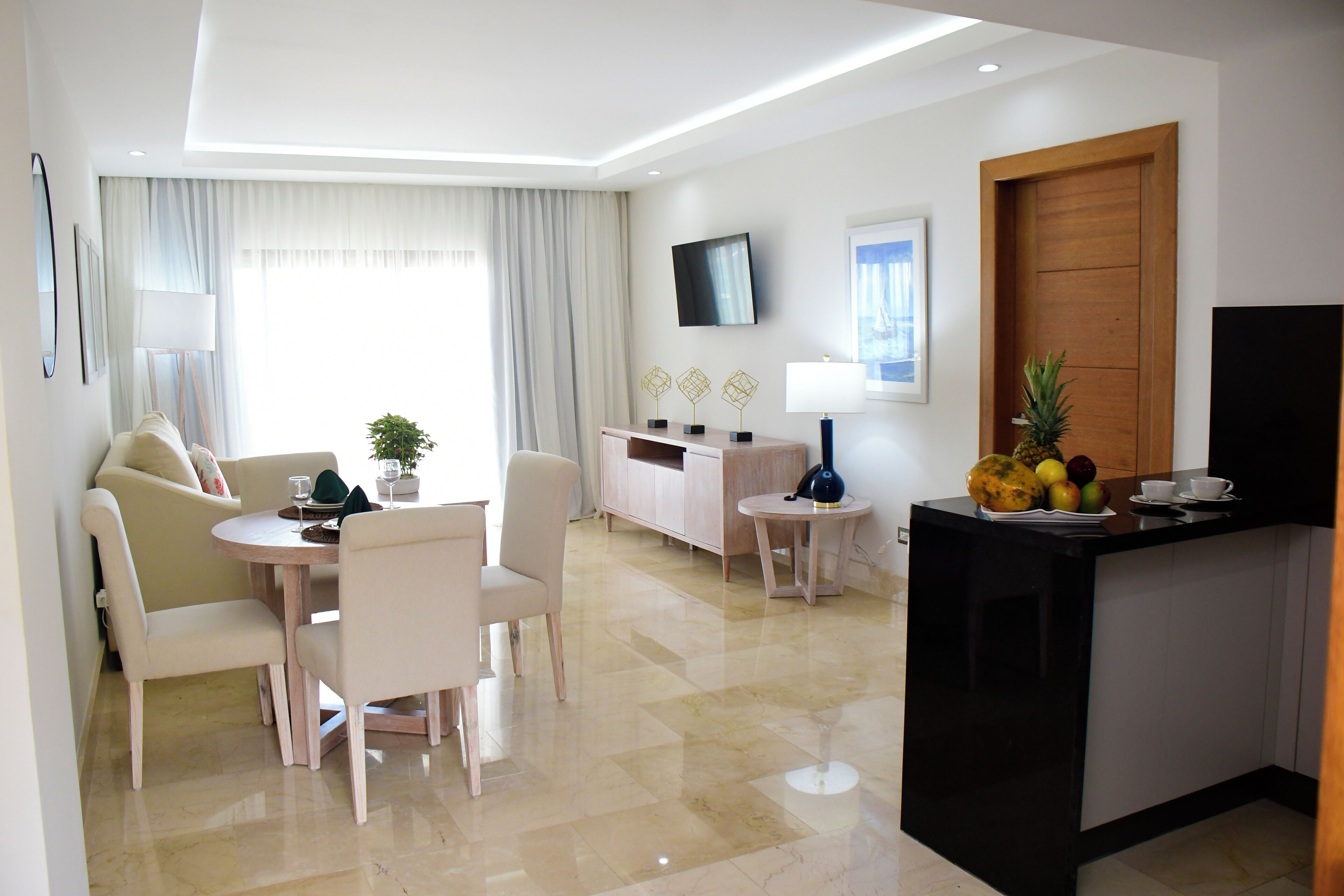 Une chambre d’hôtel moderne dotée d’un coin repas, d’un comptoir de cuisine avec des fruits, d’une télévision et d’une grande fenêtre avec des rideaux.