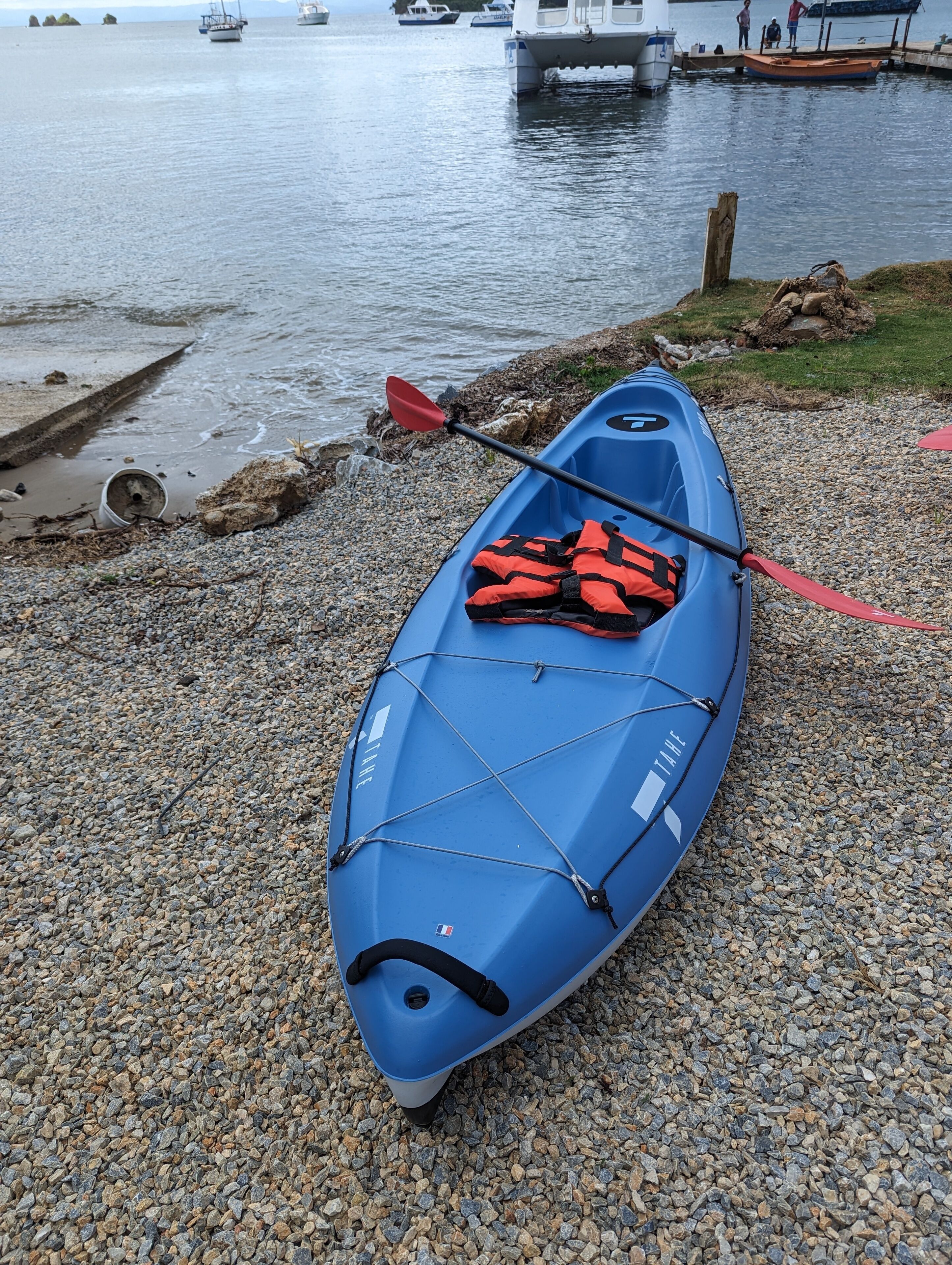 Een blauwe kajak met een reddingsvest en peddel op een kiezelstrand, aan een kalm waterlichaam.