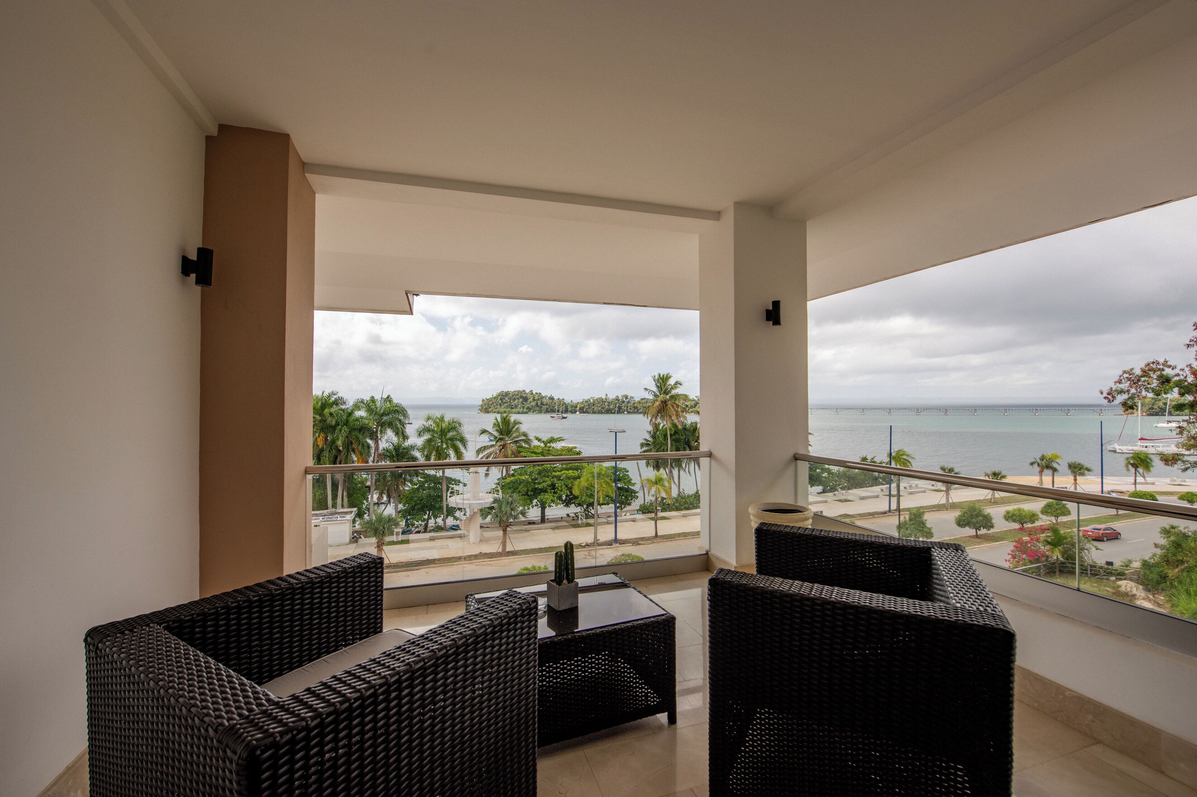 A balcony with wicker furniture, a glass door, and a view of the sea and palm trees.