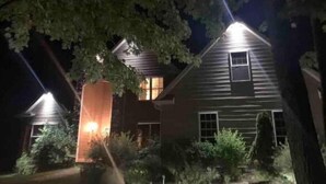 Casa de dois andares com chaminé visível e uma janela iluminada, rodeada por árvores e arbustos.
