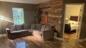 Uma sala de estar com sofá seccional, piso de madeira, uma janela grande e uma bandeira americana fixada na parede.