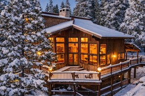 Une cabane en rondins dotée de nombreuses fenêtres, d’une terrasse et entourée d’arbres couverts de neige.