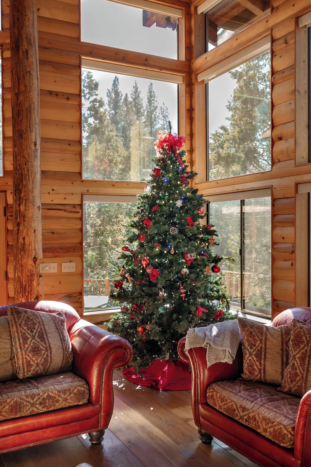 Interior acollidor d'una cabana amb una paret de fusta, grans finestres, un arbre de Nadal i sofàs de cuir.