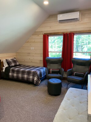 Ein gemütliches Zimmer mit einem Bett, zwei Stühlen, einem weißen Hocker und einem Fenster mit roten Vorhängen.