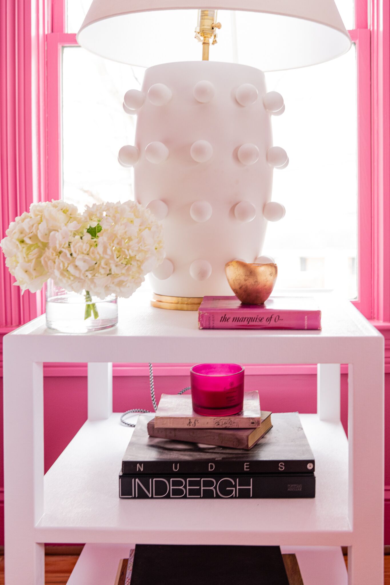A white bedside table with a lamp, a vase of flowers, and books.