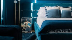 A modern bedroom with a teal color scheme, featuring a bed with blue and white bedding, a clear bedside table with books, and a large abstract painting above the bed. - The Historic Magnolia House (Greensboro)
