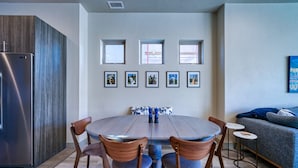 A modern kitchen with a stainless steel refrigerator, a wooden cabinet, and a dining table with chairs. There are framed pictures on the wall above the table.
