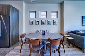 A modern kitchen with a stainless steel refrigerator, a wooden cabinet, and a dining table with chairs. There are framed pictures on the wall above the table.