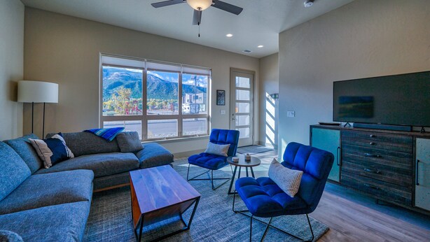 A modern living room with a sectional sofa, two blue armchairs, a coffee table, a ceiling fan, a television, and a large window offering a mountain view.