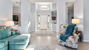 A spacious living room with a light blue sofa, a floral armchair, and a large rug. A hallway leads to a door at the end.